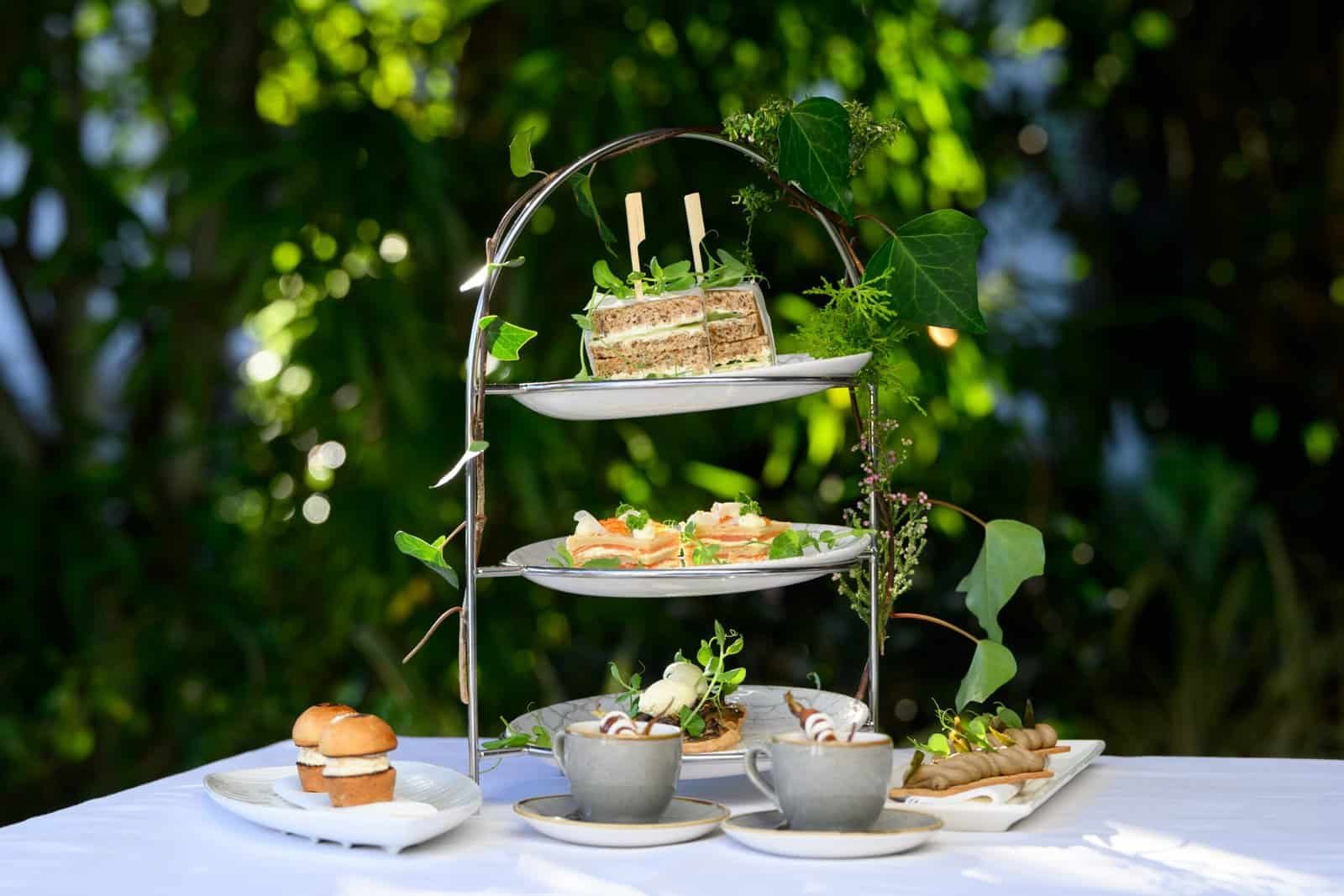 Elegant high tea assortment featuring sandwiches, scones, and desserts on a tiered stand amidst lush greenery at Palm House Boutique Hotel and Spa.