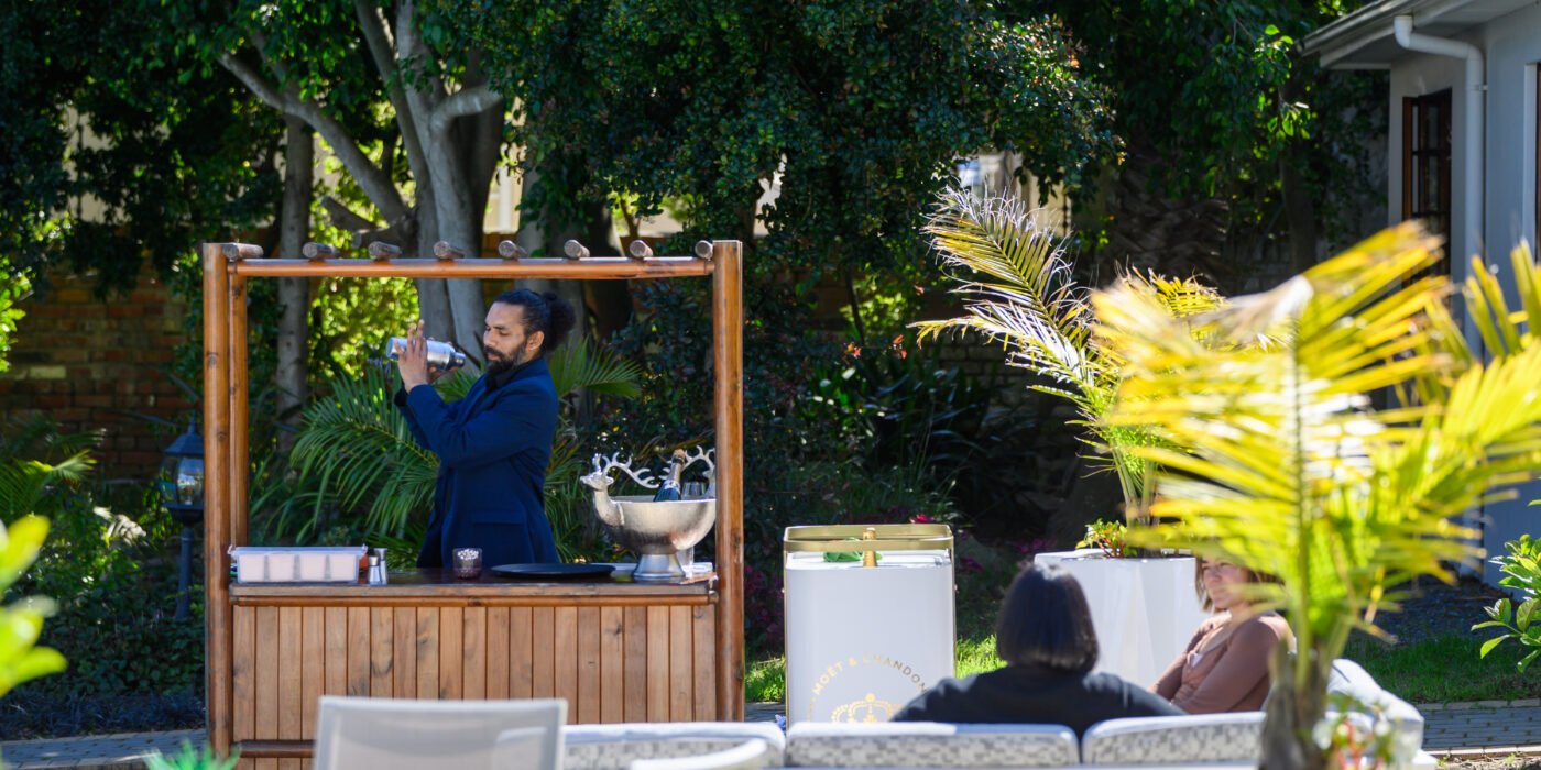 Patio area at Palm House Boutique Hotel and Spa in Johannesburg, featuring outdoor lounge, lush greenery, and a bartender preparing drinks, ideal for luxury hospitality and relaxation.