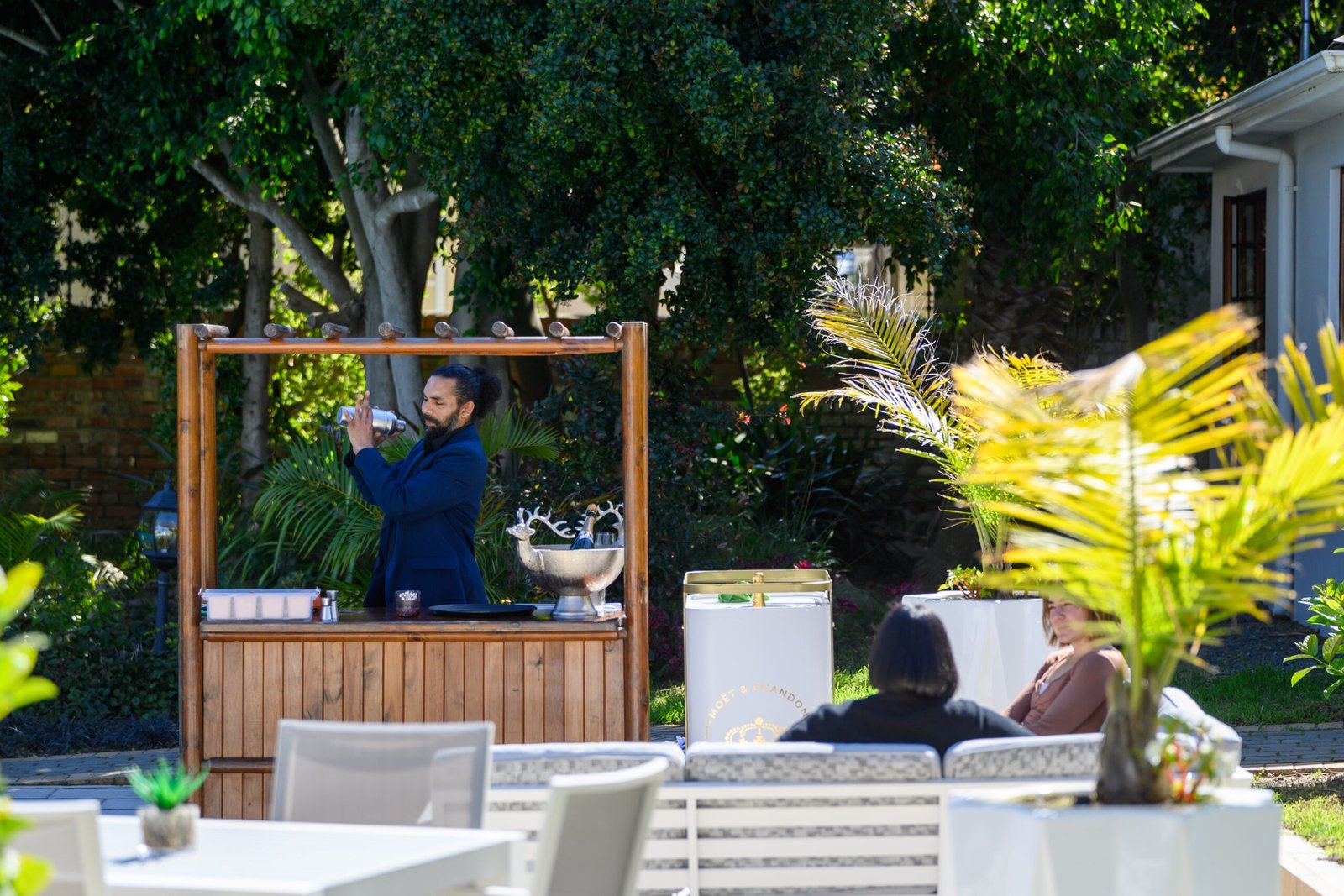 Patio area at Palm House Boutique Hotel and Spa in Johannesburg, featuring outdoor lounge, lush greenery, and a bartender preparing drinks, ideal for luxury hospitality and relaxation.