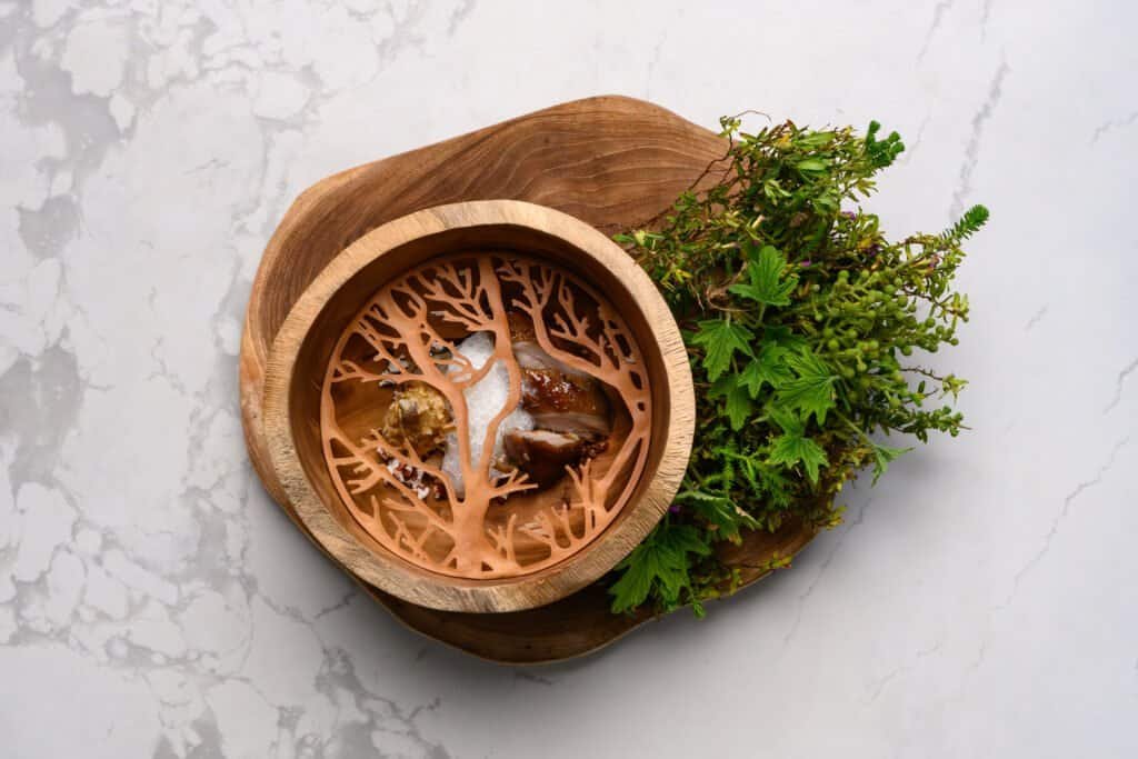 Delicate wooden presentation bowl with rustic meats and sauces, accompanied by fresh green herbs, on a white marble surface at Palm House Boutique Hotel and Spa in South Africa.